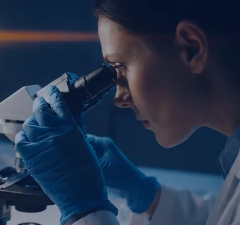 A close-up view of a person, likely a scientist or researcher, looking into a microscope while wearing blue gloves and working in a dimly lit laboratory setting