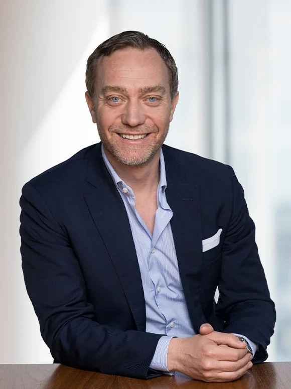 The image shows a smiling middle-aged man with dark hair and blue eyes, wearing a navy suit and light blue striped shirt, seated at a desk against a white background