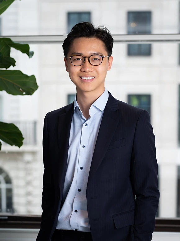 A well-dressed Asian man with glasses, wearing a black suit and a light blue shirt, smiling confidently in an office setting