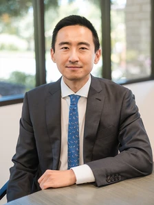The image shows James Kang, a professional-looking man in a gray suit and blue patterned tie, sitting at a table and smiling at the camera