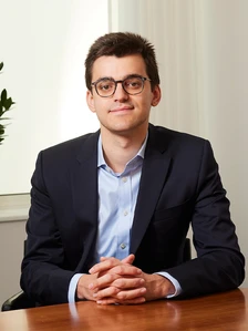 The image shows a young man with short, dark hair wearing a suit and glasses, sitting at a desk and looking directly at the camera with a friendly expression