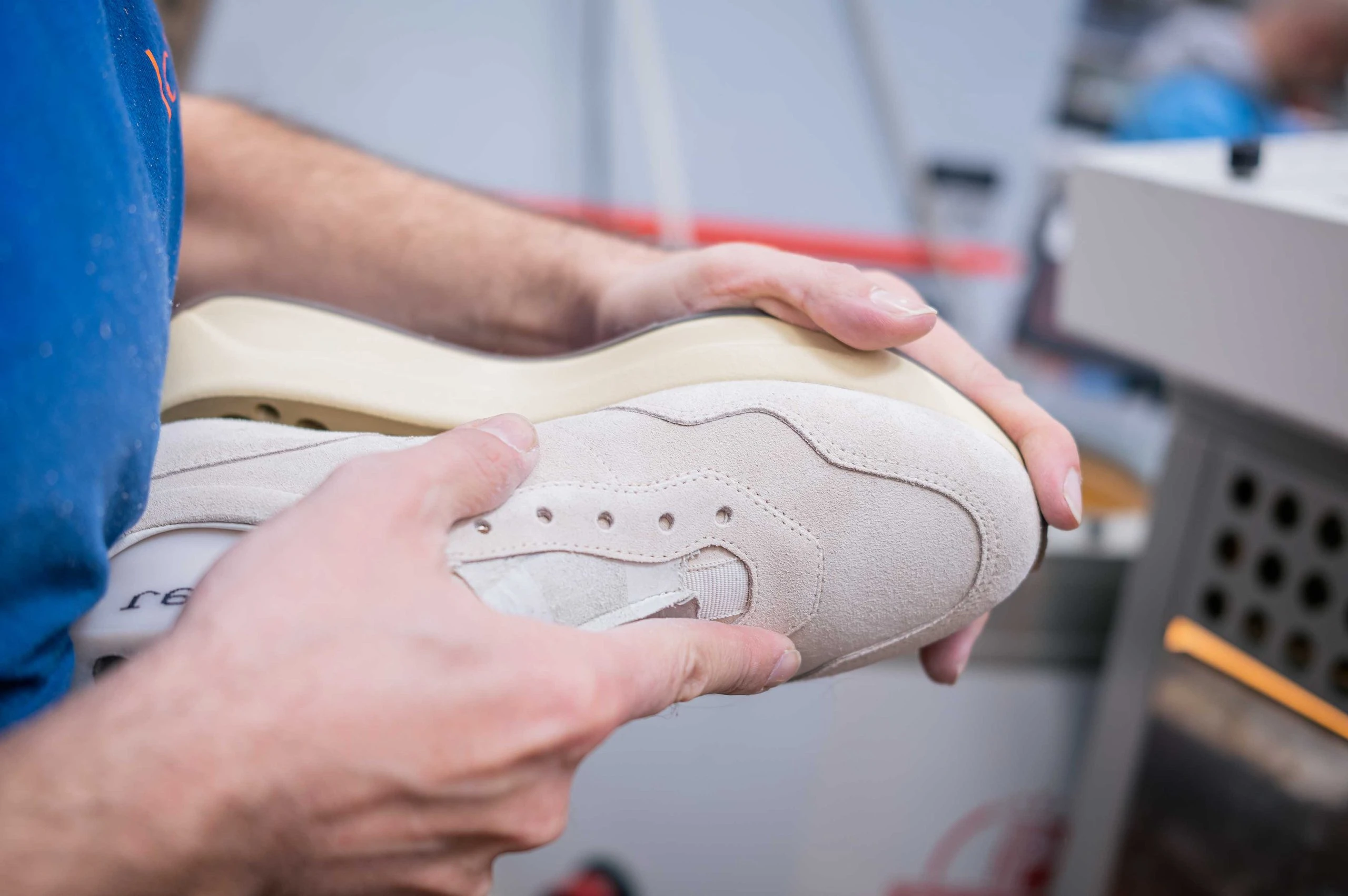 The image shows a person's hands assembling or repairing a pair of gray sneakers or shoes in what appears to be a workshop or repair shop