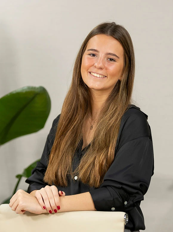 The image shows a smiling young woman with long, dark hair wearing a black top and sitting in front of a green plant