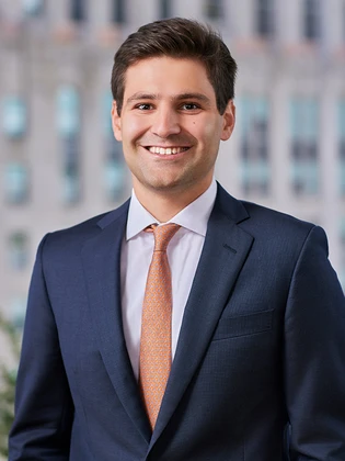 A smiling middle-aged man in a navy suit and orange patterned tie, standing against a blurred backdrop, identified as Aaron Casella