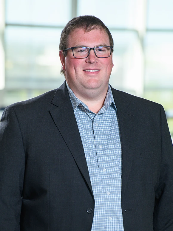 Jay Thorton, a middle-aged man wearing glasses and a checkered shirt, stands in front of a glass wall, smiling confidently