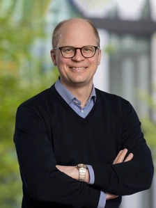 The image shows a smiling middle-aged man wearing glasses, a blue and white checkered shirt, and a black sweater, with his arms crossed, standing in front of a blurred natural background