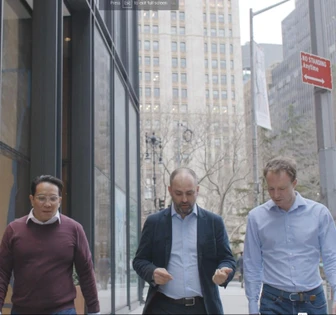 Three middle-aged men in business attire walking down a city street with skyscrapers in the background