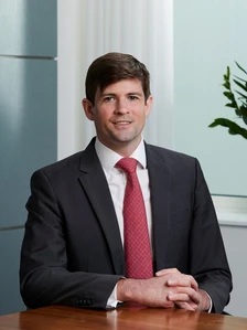 A well-dressed man with a friendly expression sitting at a desk, wearing a dark suit and red tie, appears to be Kenneth Waterson based on the image filename