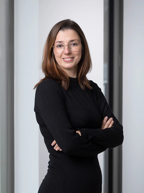 The image shows a woman with long, dark hair wearing a black sweater and round glasses, standing in front of a white background with a serious expression on her face