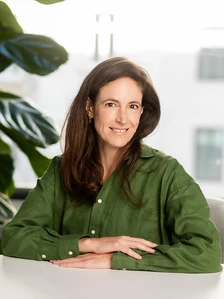 Portrait of Claire Nicoll, a smiling woman with long brown hair wearing a green jacket, sitting at a table with plants in the background
