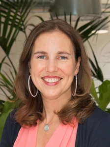 The image shows a smiling middle-aged woman with long brown hair, wearing a pink shirt and navy jacket, standing in front of lush green plants