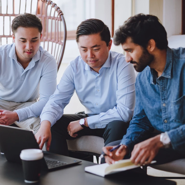Three men in business attire appear to be collaborating on a laptop during a meeting, suggesting a diverse professional setting