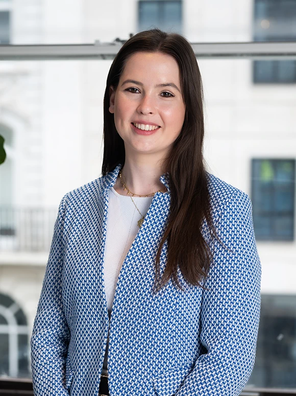 Photograph of Hannah Walsh, a business professional wearing a blue and white patterned jacket, standing in an office setting and smiling at the camera
