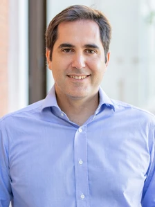 A smiling middle-aged man with short dark hair, wearing a blue dress shirt, posing for a portrait