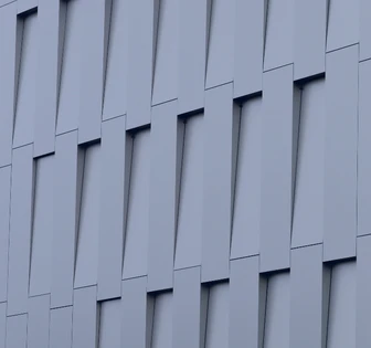 The image shows an abstract architectural facade with angled, overlapping grey panels creating a visually striking geometric pattern