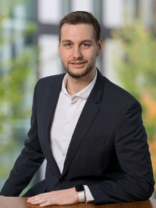 A headshot of Julian Redlich, a young man with dark hair and a beard, dressed in a black suit and white shirt, smiling and looking directly at the camera
