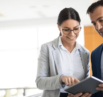 Two professionals, a woman wearing glasses and a man in a suit, review a document together while standing in an office setting