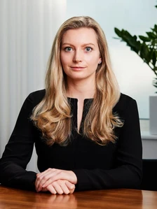 A professional woman with long blonde hair wearing a black blazer, sitting at a desk and looking directly at the camera