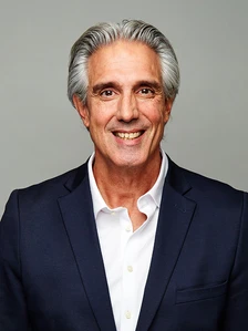 The image shows John Turner, a smiling, older man with gray hair, wearing a navy blue suit and white shirt, looking directly at the camera