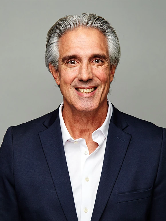The image shows John Turner, a smiling, older man with gray hair, wearing a navy blue suit and white shirt, looking directly at the camera
