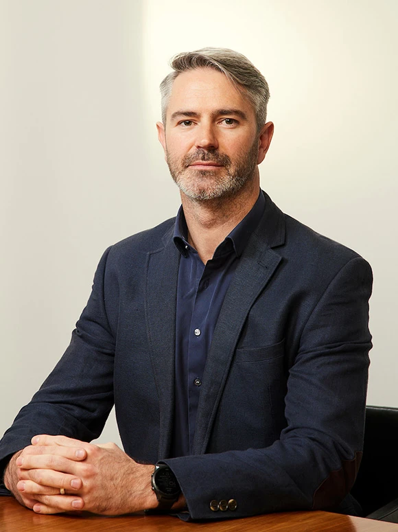 The image shows a professional headshot of Bruce Johnson, a middle-aged man with gray hair and a beard, wearing a dark suit and looking directly at the camera with a serious expression