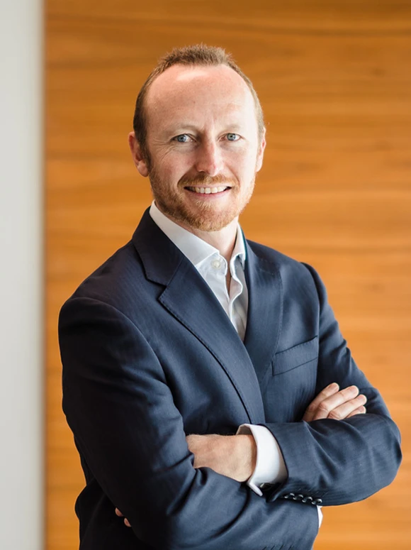 The image shows a smiling middle-aged man with a beard, wearing a dark blue suit, standing against a warm wooden background