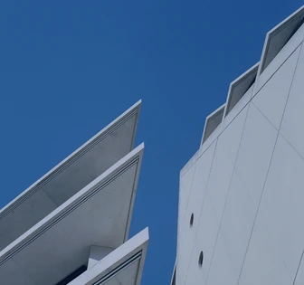 The image shows two modern, angular buildings with sharp, geometric shapes against a vibrant blue sky. The architectural design features clean lines and distinctive angular profiles