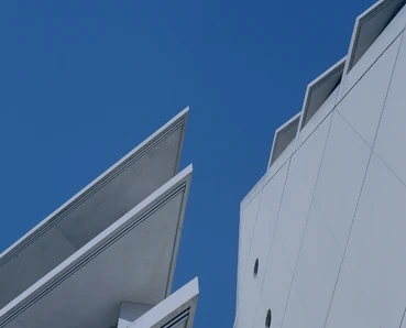 The image shows two modern, angular buildings with sharp, geometric shapes against a vibrant blue sky. The architectural design features clean lines and distinctive angular profiles