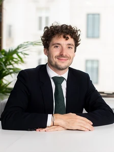 Antonin Grob, a smiling young man with curly hair, wearing a black suit and green tie, sitting at a desk in an office setting