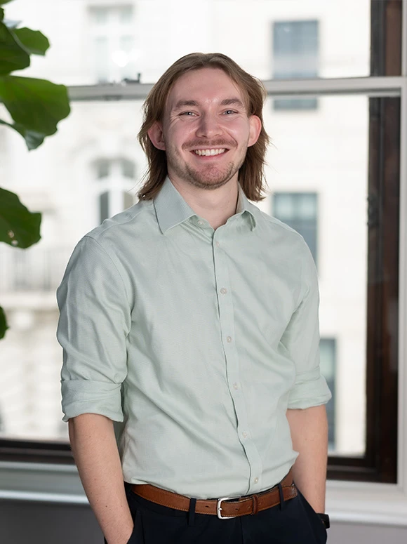 The image shows a smiling, bearded man with long brown hair, wearing a light-colored shirt, standing in front of a window