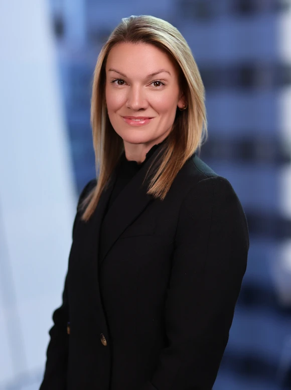 The image depicts a smiling woman with blonde hair wearing a black jacket, posing for a professional portrait against a blurred blue background