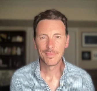 The image shows a headshot photograph of a middle-aged man with short dark hair and a friendly expression, wearing a checkered button-down shirt and looking directly at the camera
