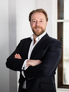 The image shows Merrill Goulding, a professional-looking man with a beard, wearing a dark suit and standing confidently with his arms crossed in front of a white wall