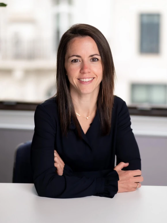 The image shows a smiling woman with long dark hair, wearing a black blazer, sitting at a desk in an office setting
