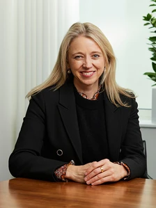 A smiling woman in a black suit and patterned blouse, seated at a desk against a curtain backdrop. The image is named "Caroline.Carr"