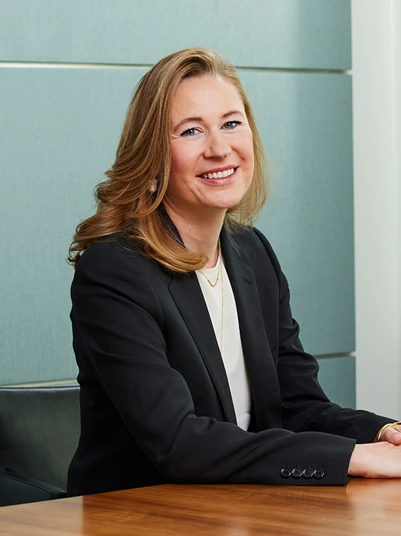 The image shows a smiling woman with long blonde hair wearing a black blazer, sitting at a desk with a light gray background