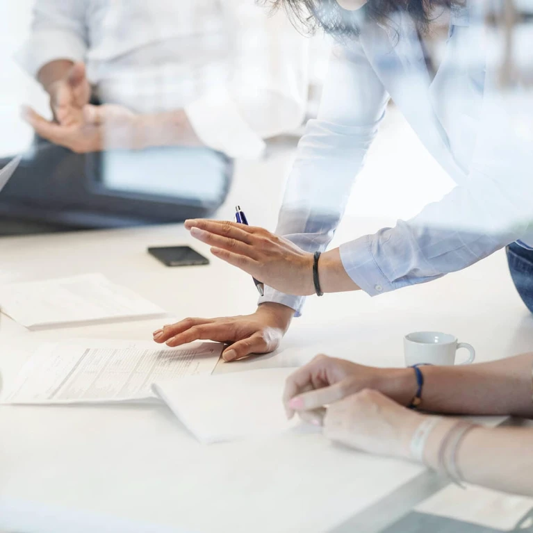 The image shows several individuals in a business setting, collaborating at a desk while using technology and office supplies