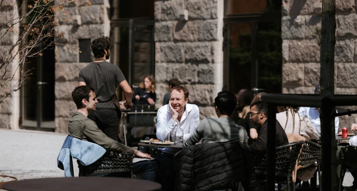 A group of people, including a man named Permira, sitting at a table and engaging in conversation at an outdoor cafe