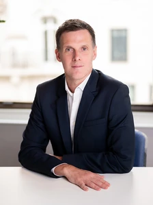 The image shows a well-dressed man in a suit, likely Pierig Melot, seated at a desk in a professional setting, with a confident and focused expression