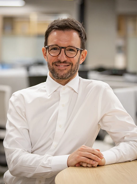The image shows Luis Fernandez, a smiling businessman wearing a white shirt and glasses, sitting at a desk in an office setting