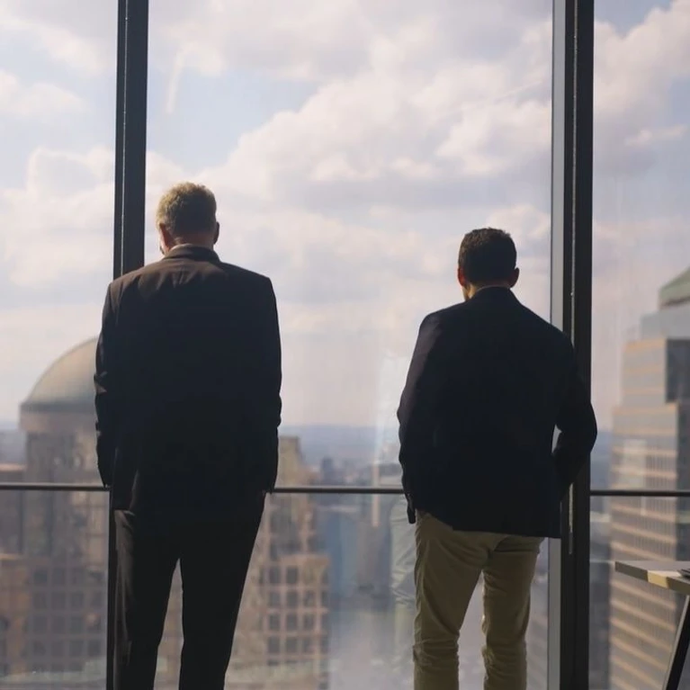 Two well-dressed individuals stand at a window, gazing out over a city skyline on a cloudy day