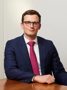 The image shows Dries Decreamer, a professional-looking young man in a suit and tie, sitting at a desk and making direct eye contact with the camera