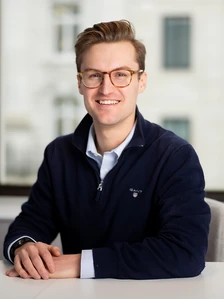 Hugo Sloper, a smiling young man with brown hair wearing glasses, a navy blue jacket, and a white shirt, posing in a professional setting