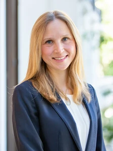 Irene Lamsaki, a middle-aged woman with long, blonde hair, smiling while wearing a navy blue blazer and white top, standing in front of a blurred background