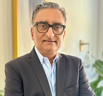 The image shows Raj Patel, a middle-aged man wearing a grey suit and black-framed glasses, standing in a professional setting with a warm expression