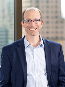 A smiling middle-aged man with gray hair, wearing glasses and a blue suit, stands in front of a glass window with a city skyline in the background