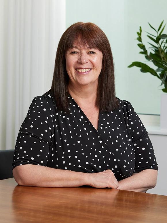 Tina Grimshaw, a smiling middle-aged woman, sits at a desk in a polished office setting, wearing a black-and-white polka dot blouse