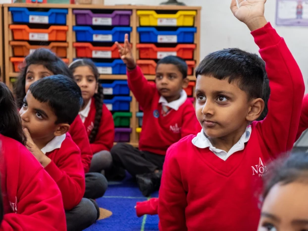 The image shows a group of young students wearing red uniforms, sitting on the floor of a classroom with colorful storage shelves behind them. One student is raising their hand