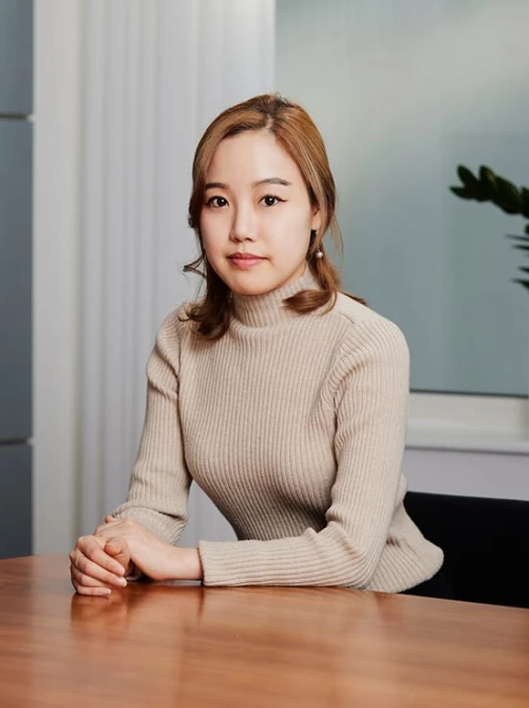 The image shows a young woman with long brown hair, wearing a beige turtleneck sweater, sitting at a table with her arms folded, looking directly at the camera with a serious expression