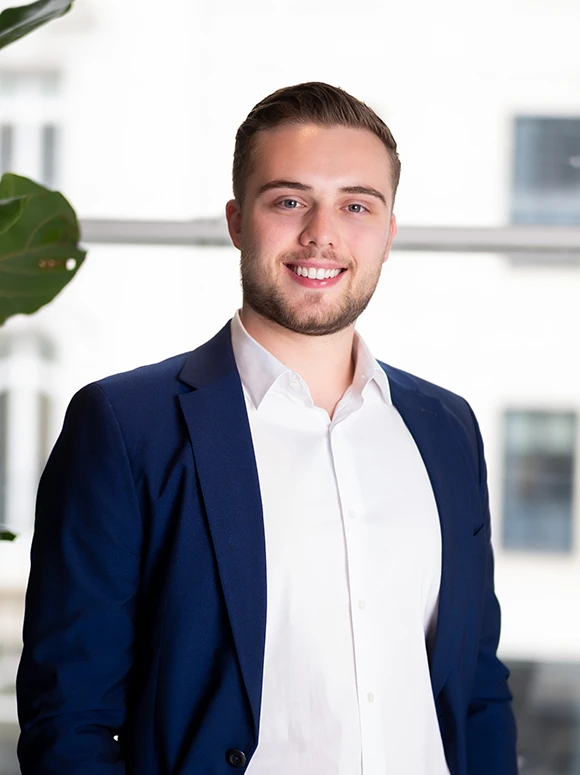 The image shows a smiling young man wearing a navy blue suit and white shirt, standing in a professional office setting